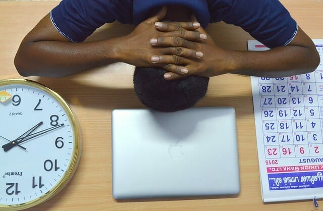 一个过度疲劳的男人把头放在桌子上，旁边是他的笔记本电脑、日历和时钟